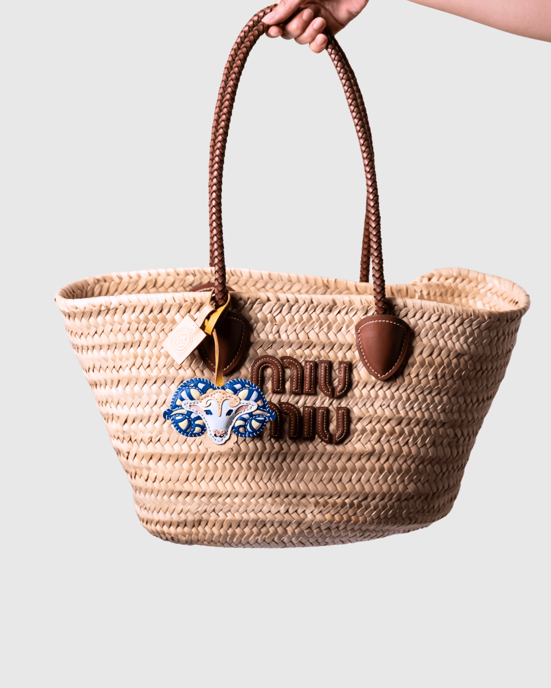 Hand holding a woven straw tote bag with brown leather handles.
