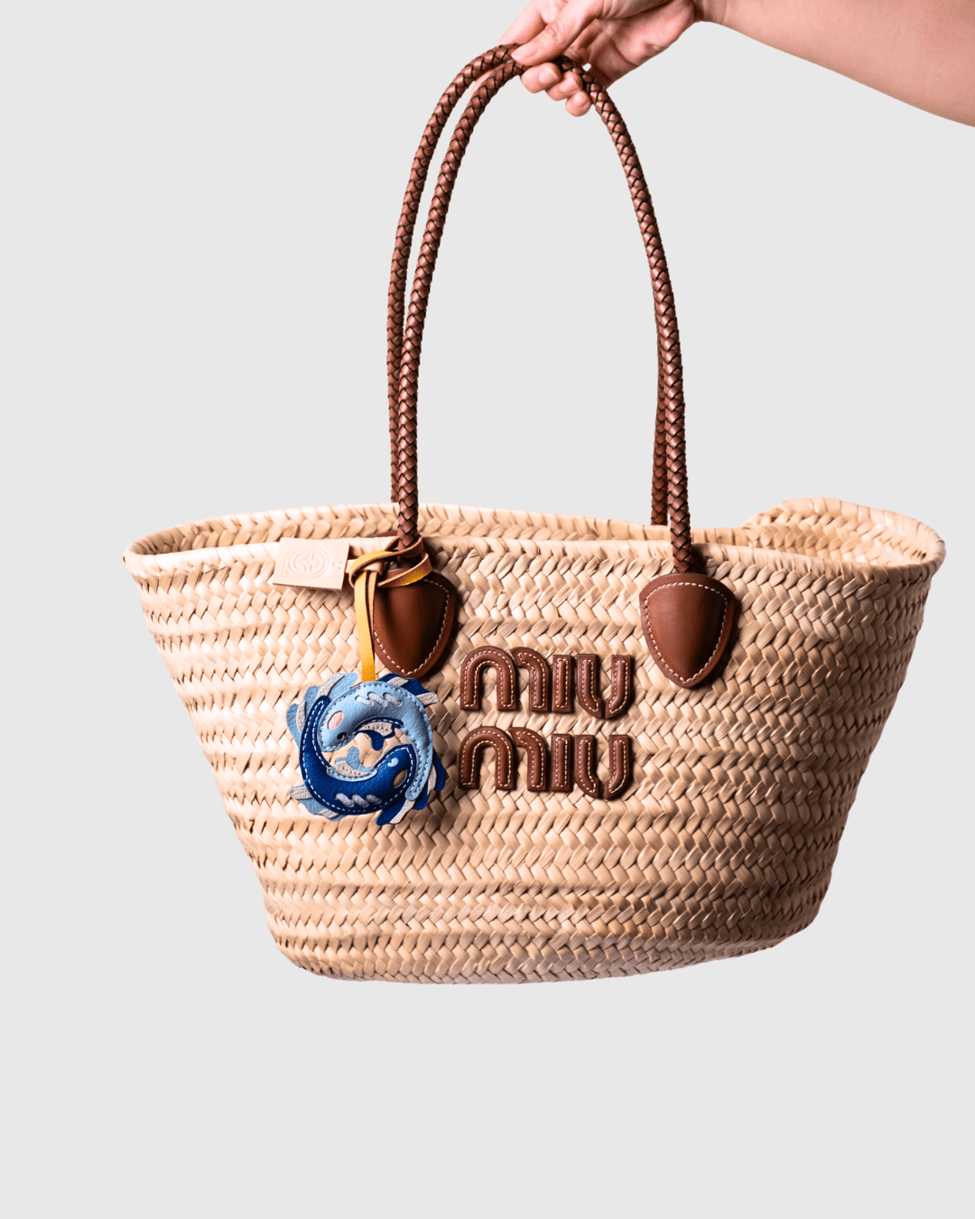 Woven straw handbag with leather handles and logo, held by a hand.
