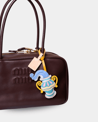 A close-up of a dark brown leather handbag with gold-tone hardware, featuring an embroidered patch of a blue and yellow jester hat hanging from a pink tag.
