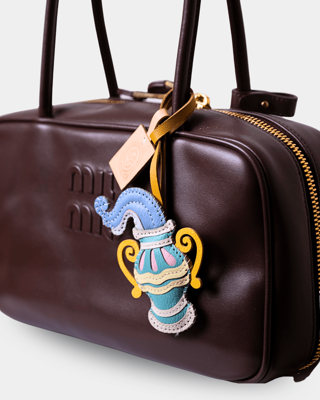 A close-up of a dark brown leather handbag with gold-tone hardware, featuring a colorful embroidered patch shaped like a teapot hanging from a gold ribbon.