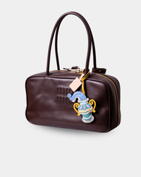 A dark brown leather handbag with a top handle, gold-tone zipper, and a decorative hanging charm shaped like a blue and gold teapot, shown on a light gray background.
