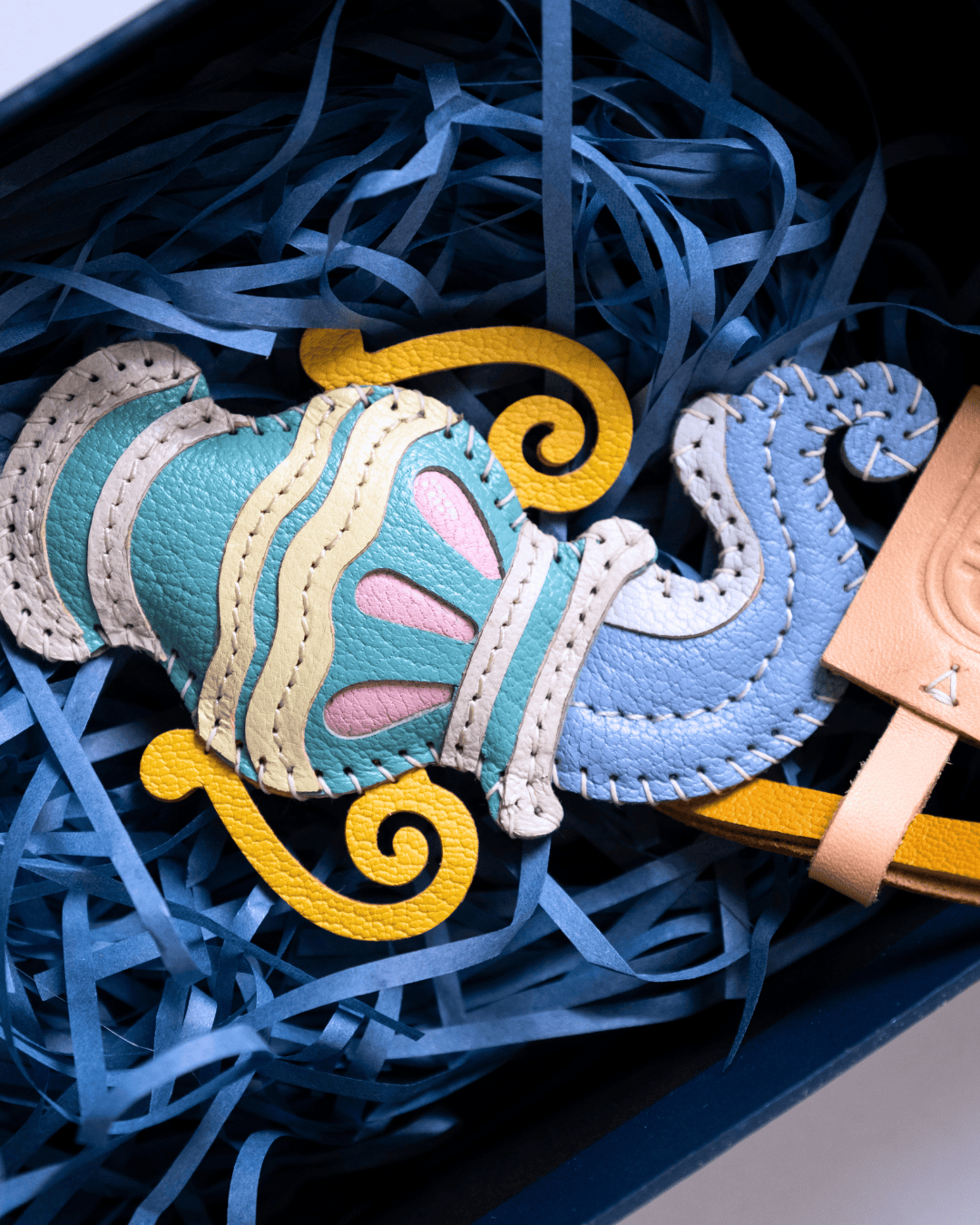 A colorful handmade leather ornament or decorative piece shaped like a stylized character, displayed inside a dark box filled with blue shredded paper. The ornament features light blue, mint green, cream, yellow, and pink sections with visible stitching.