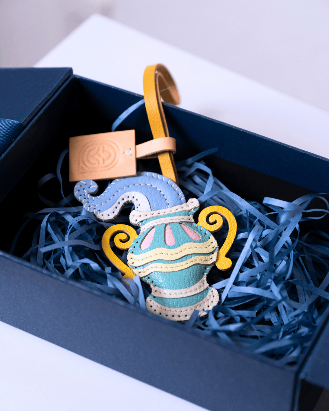 A decorative leather pouch ornament shaped like a stylized vase, displayed inside a dark blue gift box filled with blue paper shred. The pouch is light blue with yellow handles, cream trim, and small pink accents, with a tan leather tag attached.