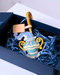 A decorative leather pouch ornament shaped like a stylized vase, displayed inside a dark blue gift box filled with blue paper shred. The pouch is light blue with yellow handles, cream trim, and small pink accents, with a tan leather tag attached.