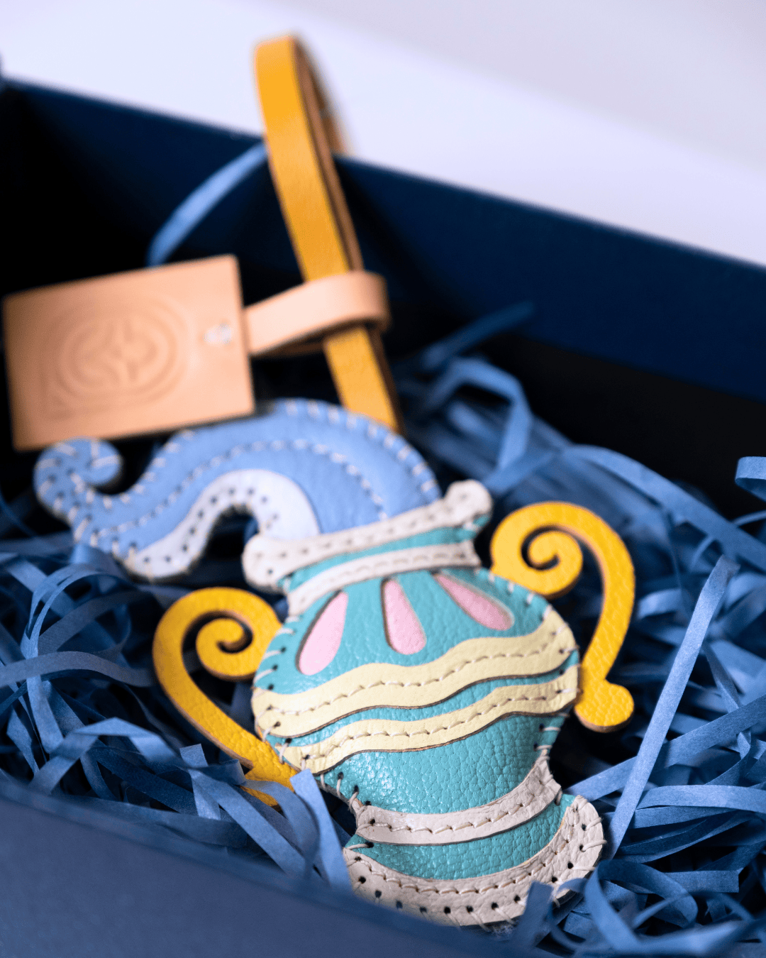 A colorful handmade leather ornament shaped like a stylized vase, displayed inside a dark blue gift box filled with blue shredded paper. The ornament features light blue, yellow, pink, and cream leather pieces, with a tan leather tag attached.