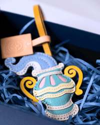 A colorful handmade leather ornament shaped like a stylized vase, displayed inside a dark blue gift box filled with blue shredded paper. The ornament features light blue, yellow, pink, and cream leather pieces, with a tan leather tag attached.