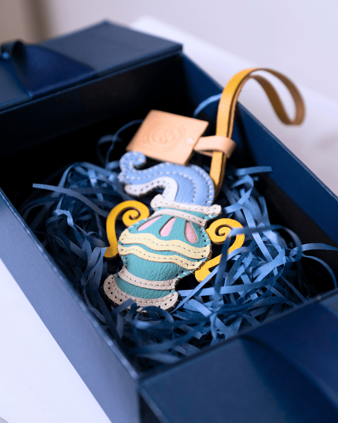 A colorful embroidered seahorse-shaped ornament resting inside a dark navy gift box filled with blue shredded paper. The ornament features a light blue seahorse body with yellow, white, and pink embroidered details, attached to a tan leather strap.