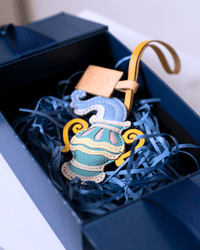 A colorful embroidered seahorse-shaped ornament resting inside a dark navy gift box filled with blue shredded paper. The ornament features a light blue seahorse body with yellow, white, and pink embroidered details, attached to a tan leather strap.