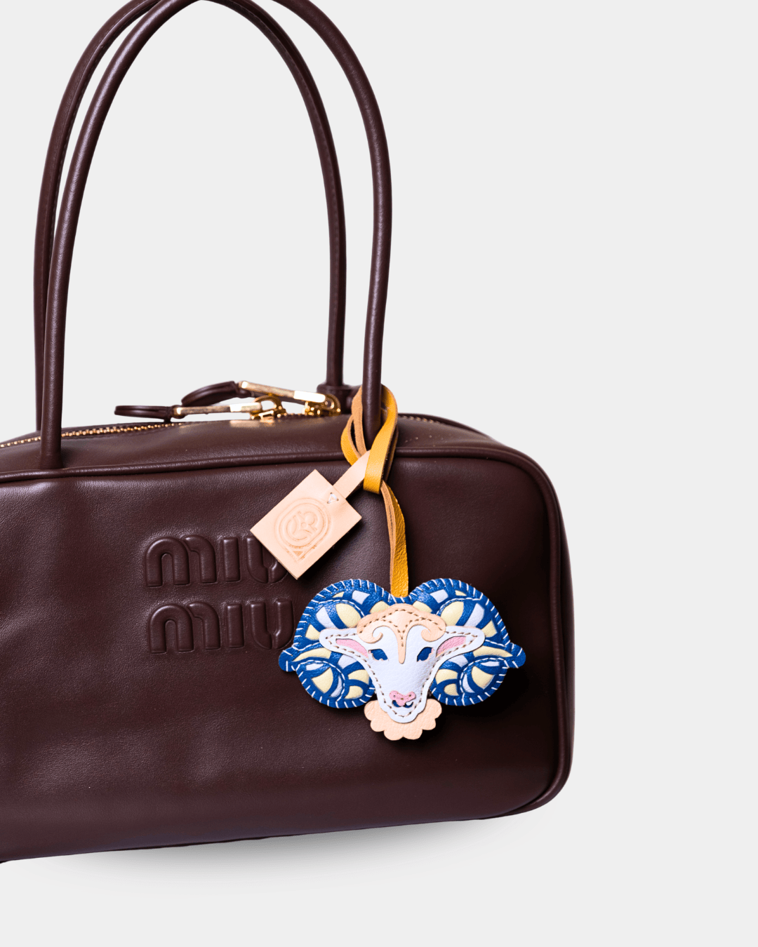 A close-up of a dark brown leather Miu Miu handbag with gold-tone hardware, featuring an embroidered blue and white goat motif charm hanging from a yellow strap and a small beige brand tag.