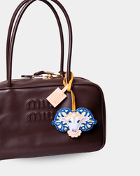 A close-up of a dark brown leather Miu Miu handbag with gold-tone hardware, featuring an embroidered blue and white goat motif charm hanging from a yellow strap and a small beige brand tag.