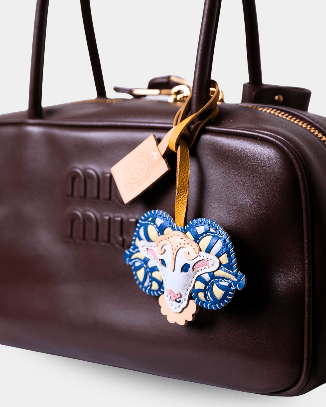 A close-up of a dark brown leather Miu Miu handbag featuring embossed logo lettering, gold-tone hardware, and a decorative blue and white embroidered sheep charm hanging from a tan leather strap.