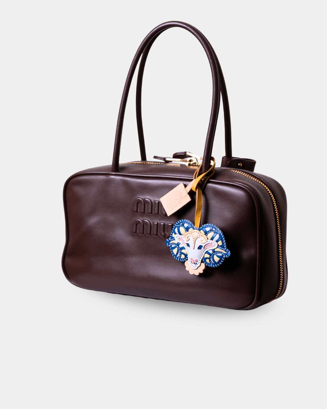 A dark brown leather Miu Miu handbag with gold-tone hardware, featuring an embossed logo on the front, dual top handles, and a decorative blue and white embroidered heart-shaped charm.