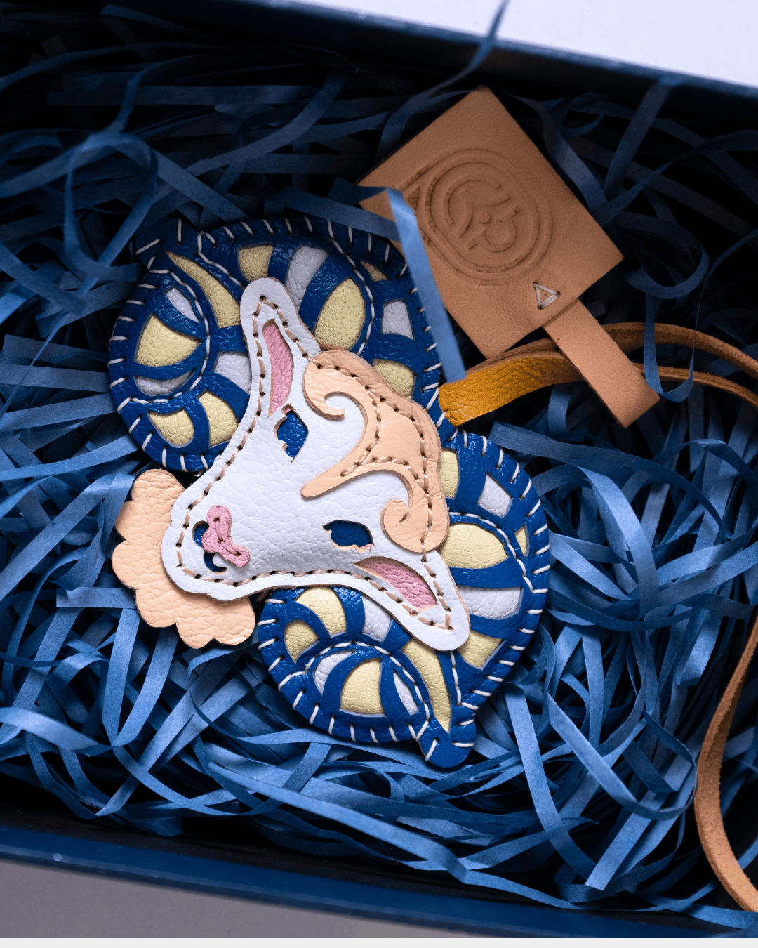 A Gucci leather accessory, likely a bag charm or key fob, featuring a stylized horse head motif in beige, white, and pink, layered over a blue and cream patterned Gucci wheel design. The item is presented in a box filled with blue paper shred alongside a tan Gucci leather tag.