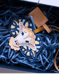 A Gucci leather accessory, likely a bag charm or key fob, featuring a stylized horse head motif in beige, white, and pink, layered over a blue and cream patterned Gucci wheel design. The item is presented in a box filled with blue paper shred alongside a tan Gucci leather tag.