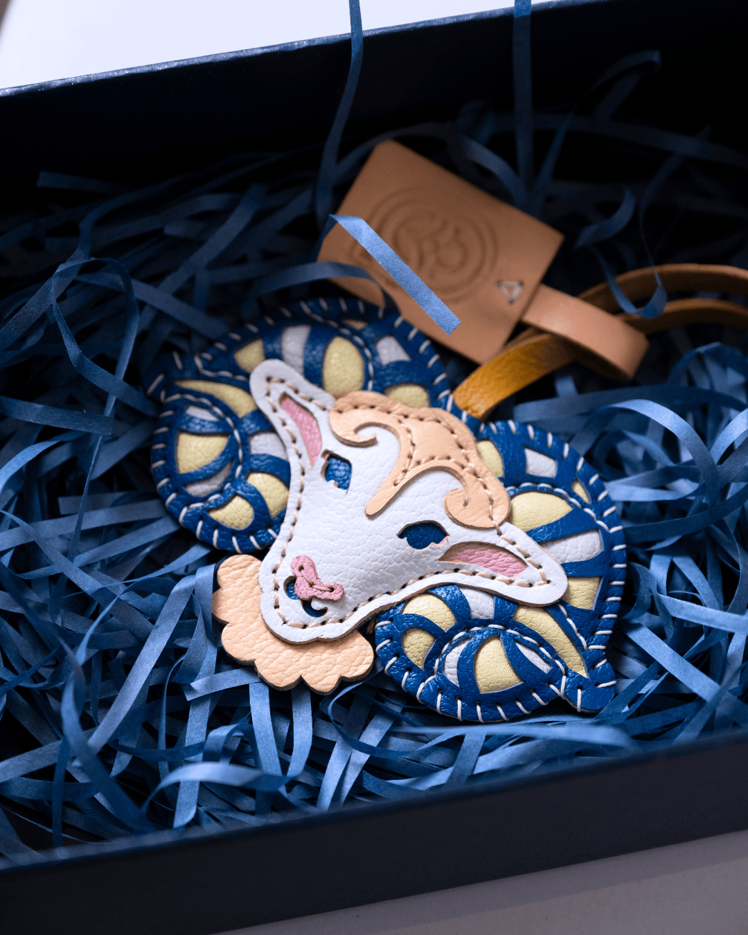 A handmade sheep-shaped leather ornament with blue and cream embroidered patterns, nestled in blue paper shred inside a black gift box with a leather tag.
