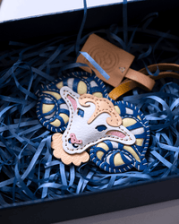 A handmade sheep-shaped leather ornament with blue and cream embroidered patterns, nestled in blue paper shred inside a black gift box with a leather tag.