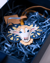 A handmade felt sheep ornament with a cream face, pink nose and ears, and blue patterned horns, nestled in blue shredded paper inside a dark gift box.
