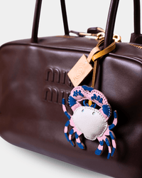 A close-up of a dark brown leather Miu Miu handbag featuring an embossed logo, gold-tone hardware, and a colorful crab-shaped leather charm in pink, blue, and white.