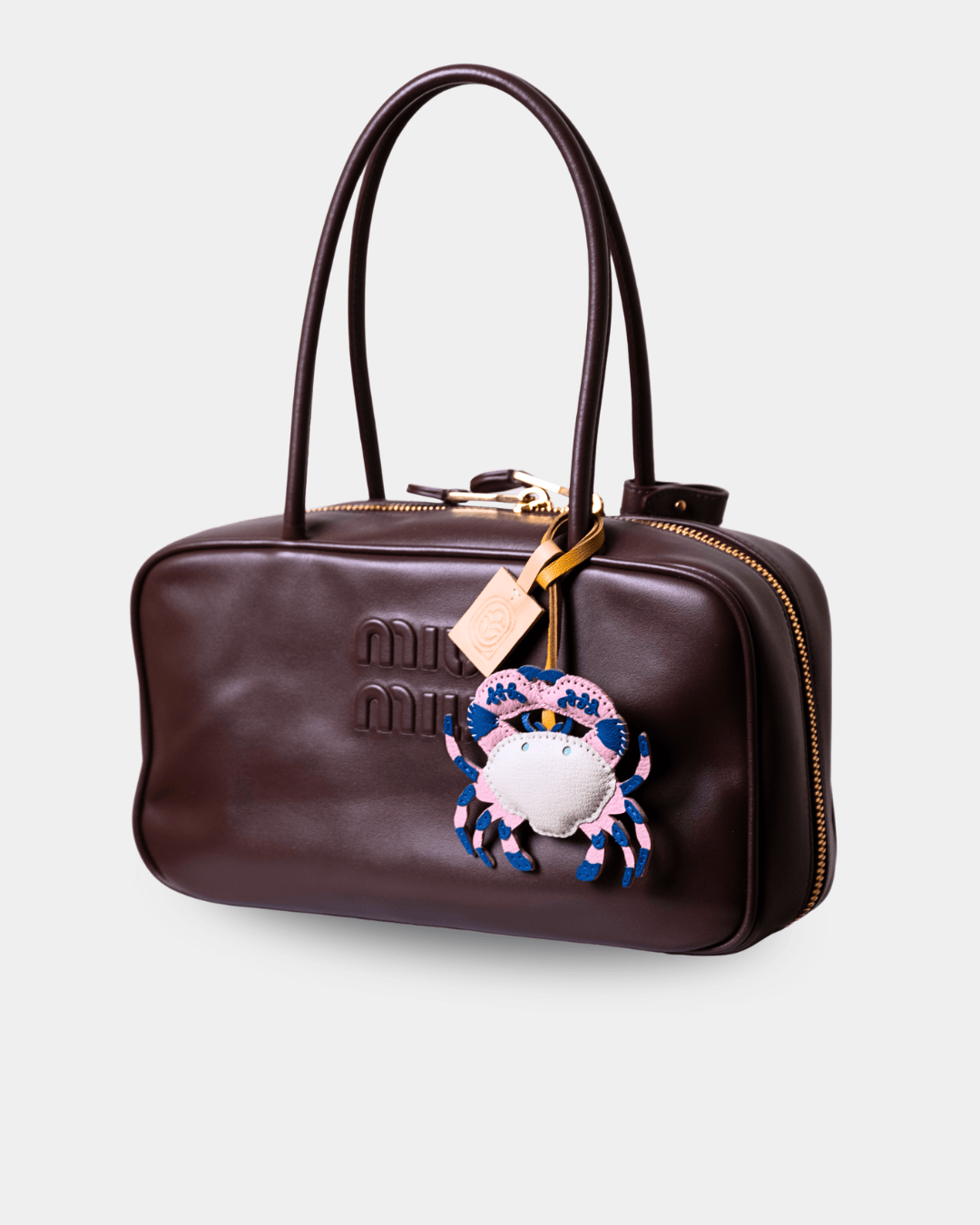 A dark brown leather Miu Miu handbag with gold-tone hardware, featuring the brand's signature embossed logo on the front, a top handle, and a decorative embroidered crab charm.