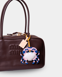 A close-up of a dark brown leather handbag with gold-tone hardware, featuring an embossed Miu Miu logo and a decorative embroidered crab charm in pink, blue, and white hanging from the handle.
