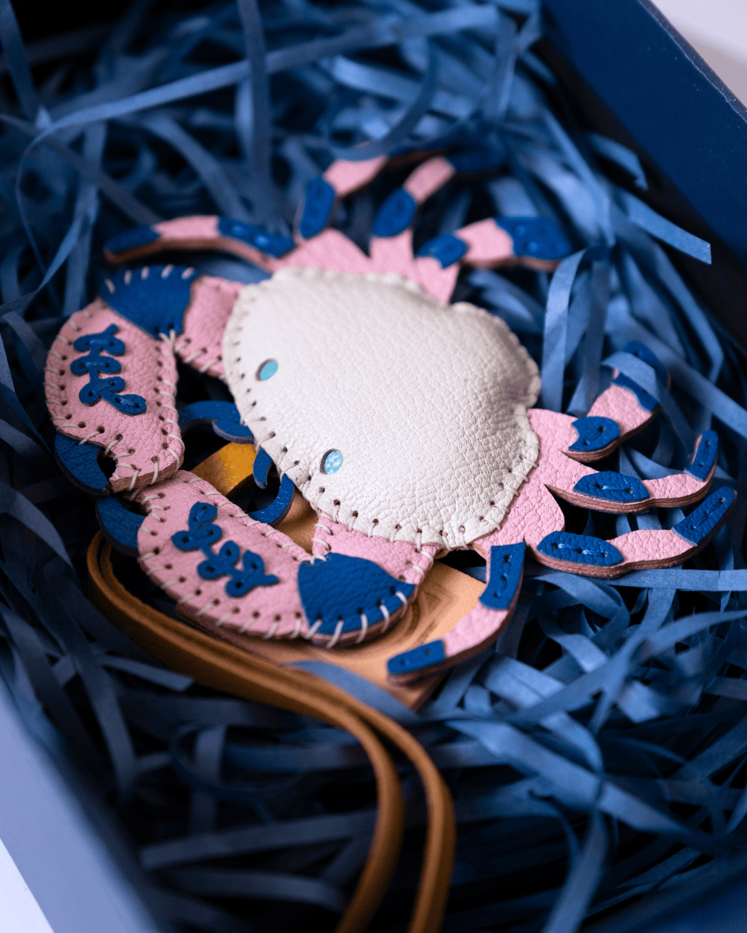 A handmade leather crab-shaped accessory, likely a belt buckle or charm, featuring a cream-colored center and pink and blue textured legs, nestled in blue paper shred inside a box.