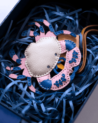 A handmade crab-shaped leather charm or accessory with a cream center, pink and blue embroidered legs, and a tan strap, nestled in blue shredded paper inside a dark box.