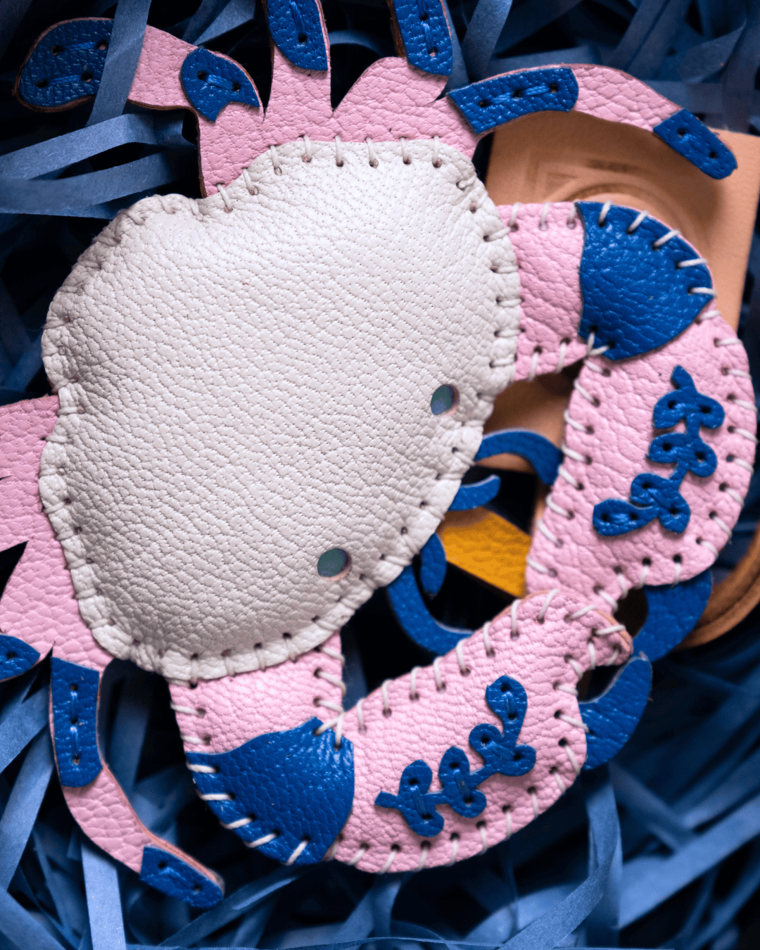 A close-up of a handmade leather crab ornament or accessory in pink and blue, layered over a beige leather mask, set against a background of blue paper shred.