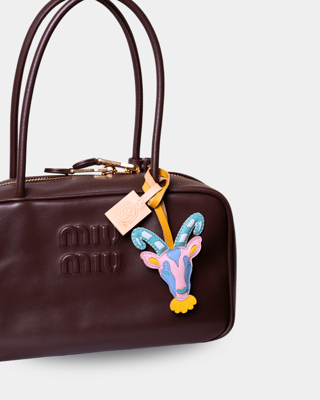 A close-up of a dark brown Miu Miu leather handbag featuring a colorful embroidered goat head charm and a branded hangtag on a light gray background.