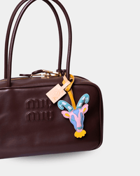 A close-up of a dark brown Miu Miu leather handbag featuring a colorful embroidered goat head charm and a branded hangtag on a light gray background.
