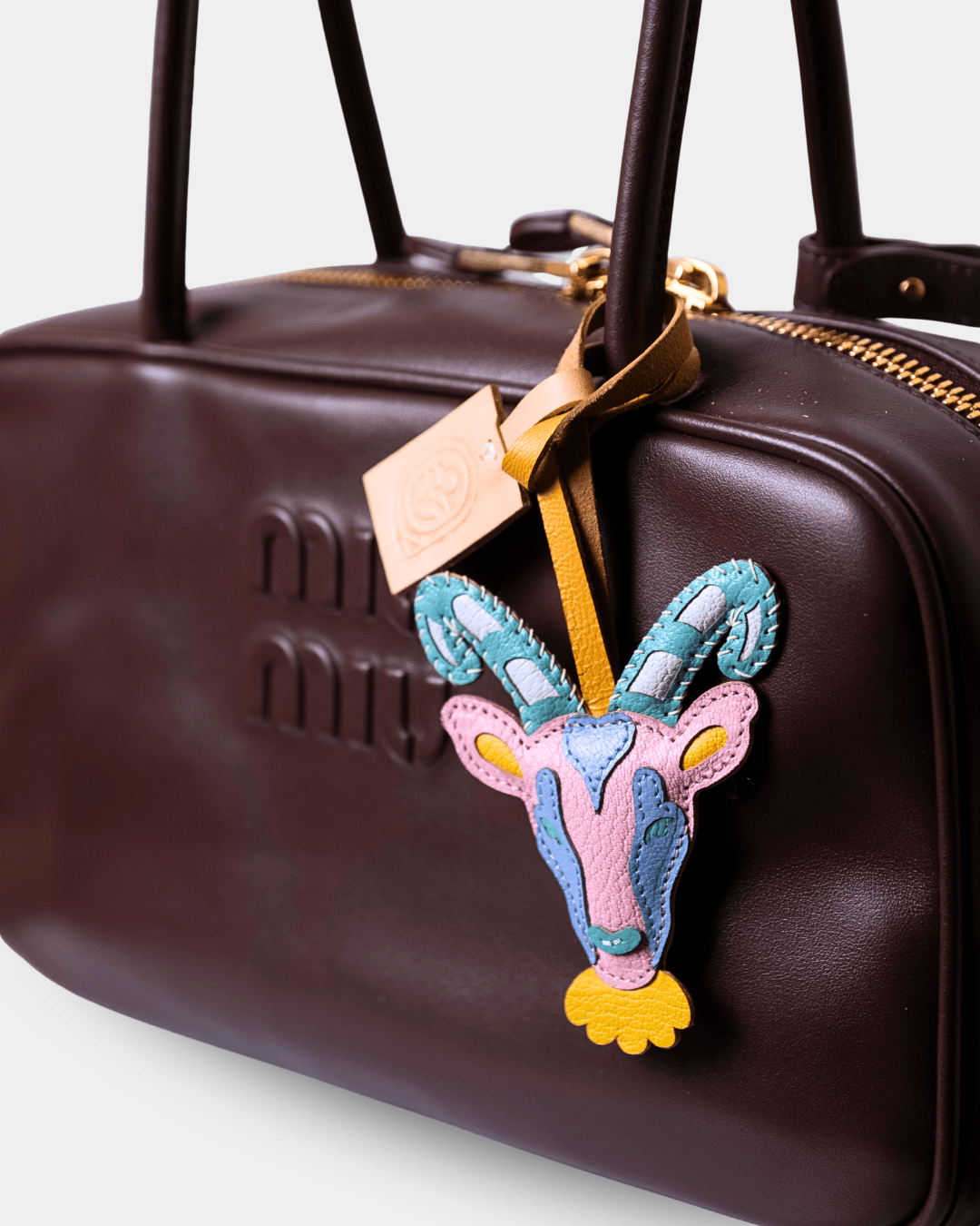 A close-up of a dark brown leather Miu Miu handbag featuring a colorful embroidered goat head charm on a gold ribbon, with the embossed Miu Miu logo visible on the front.