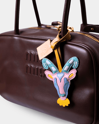 A close-up of a dark brown leather Miu Miu handbag featuring a colorful embroidered goat head charm on a gold ribbon, with the embossed Miu Miu logo visible on the front.