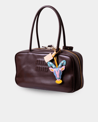 A dark brown leather Miu Miu handbag with a colorful embroidered goat head patch and a hanging beige paper tag, shown on a light gray background.