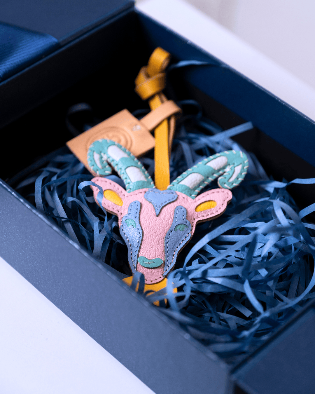 A colorful handmade leather goat head ornament with blue-green, pink, and yellow patches, nestled in blue shredded paper inside a dark navy gift box.