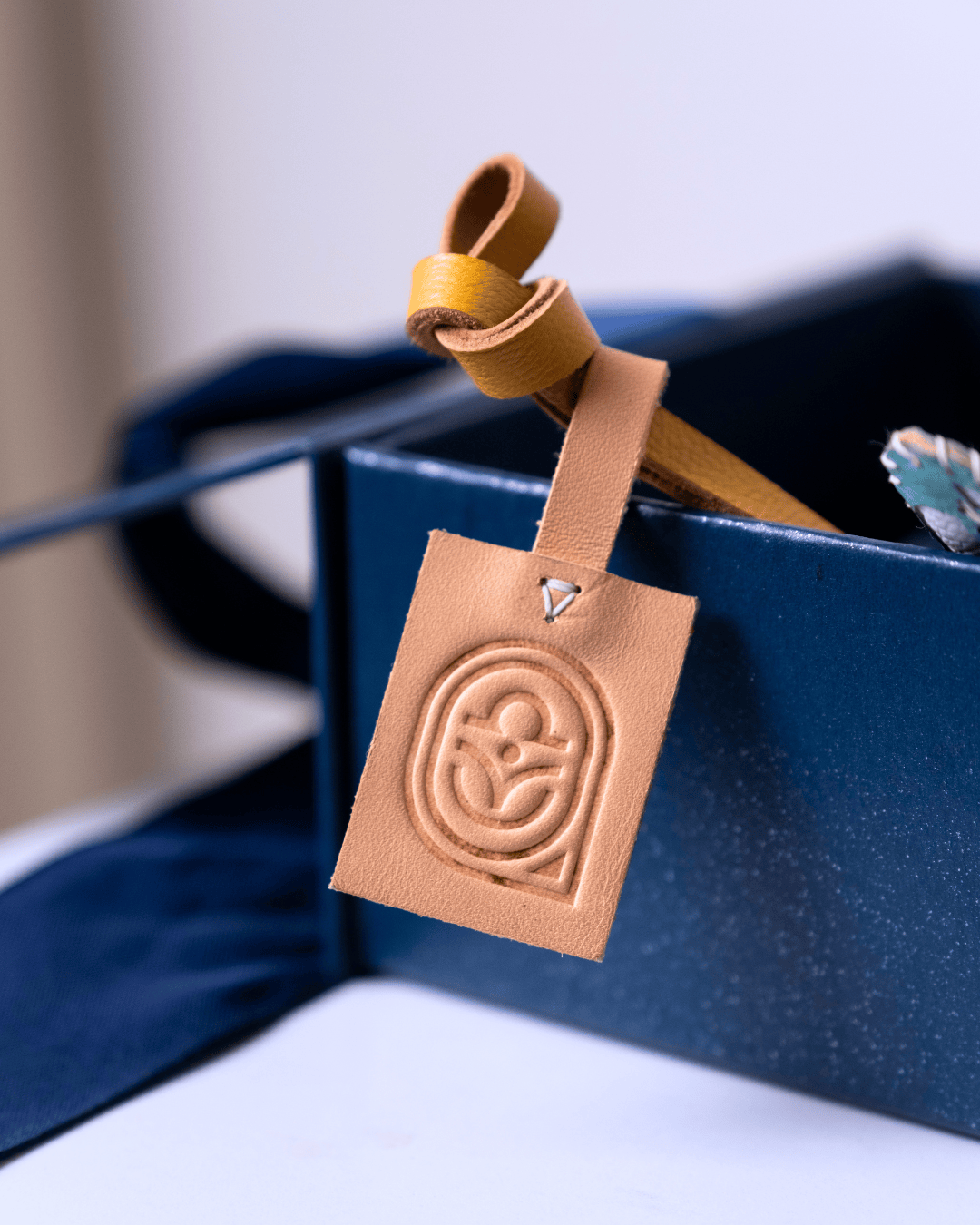 A tan leather luggage tag with an embossed circular logo, attached to a matching tan leather strap and resting inside a dark blue box.