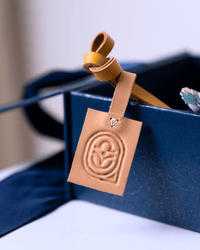 A tan leather luggage tag with an embossed circular logo, attached to a matching tan leather strap and resting inside a dark blue box.