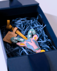 A colorful handmade leather patch shaped like a deer head, featuring pink, blue, and green panels, nestled in blue shredded paper inside a dark navy gift box. A tan leather tag with a circular logo is attached.