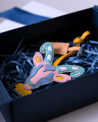 A colorful handmade leather patch shaped like a stylized deer head with antlers, displayed inside a black gift box filled with blue shredded paper.