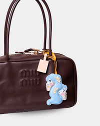 A close-up of a dark brown Miu Miu handbag featuring an embroidered light blue bunny charm with gold accents and a pink brand tag.