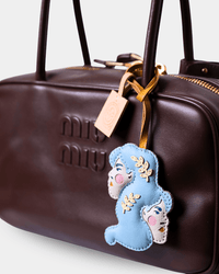A close-up of a dark brown leather Miu Miu handbag featuring embossed branding and a decorative hanging charm shaped like a blue ghost with embroidered details.
