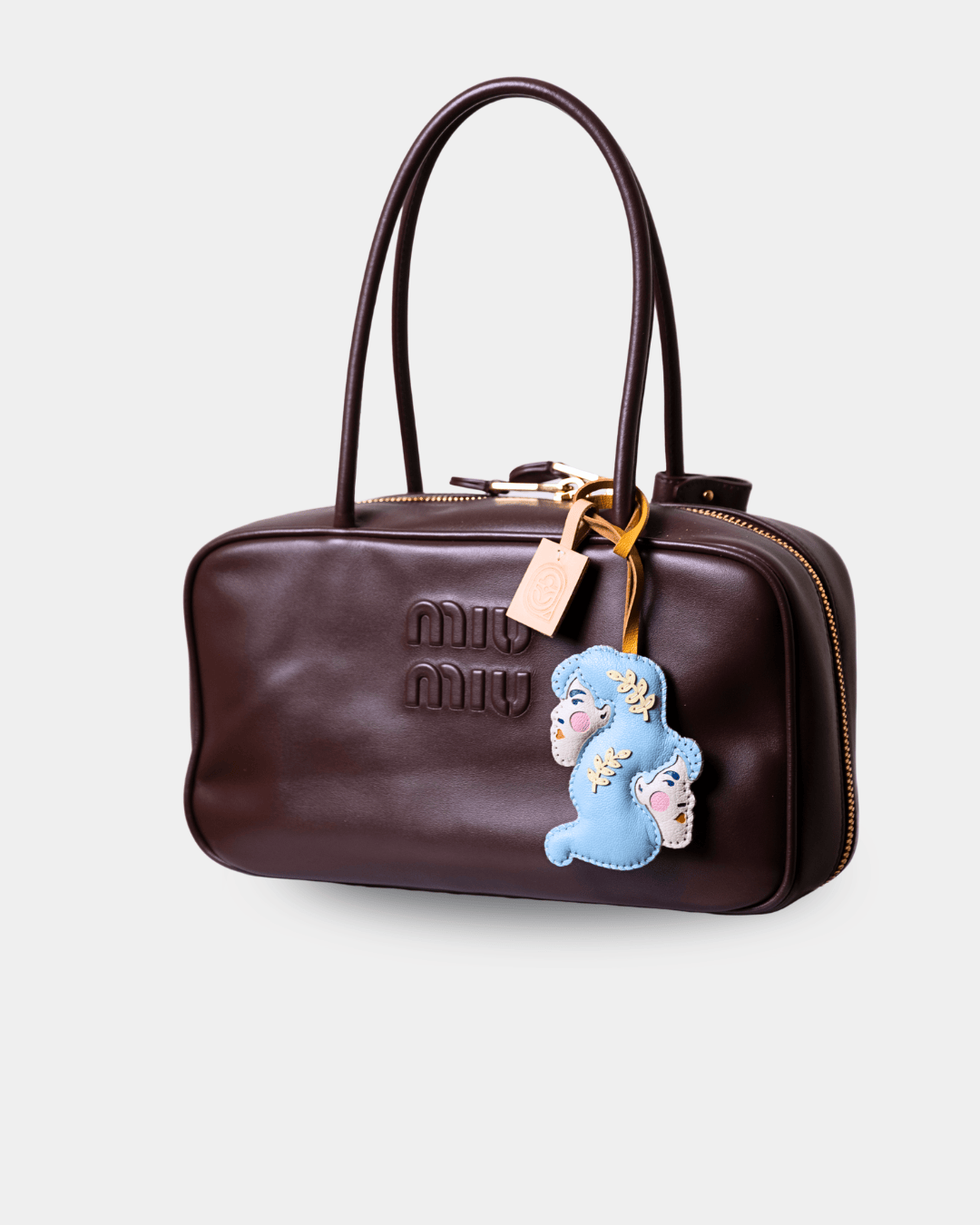 A dark brown leather Miu Miu handbag with a top handle, gold-tone zipper, and a blue plush bear charm with embroidered faces.