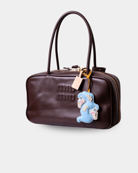 A dark brown leather Miu Miu handbag with a top handle, gold-tone zipper, and a blue plush bear charm with embroidered faces.