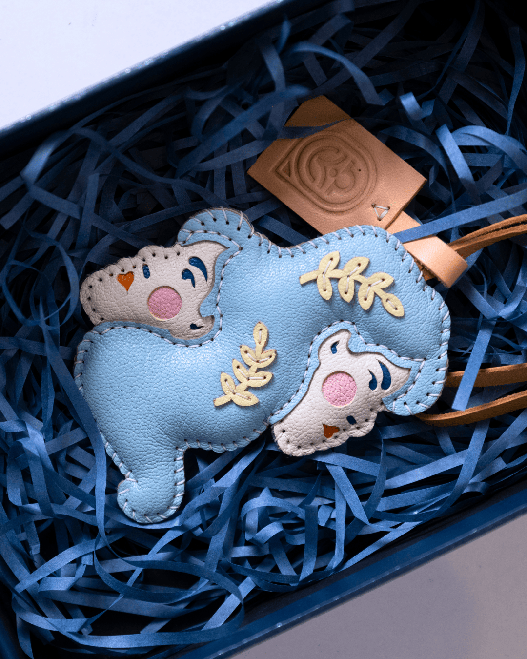 A light blue and cream faux leather bear-shaped charm with embroidered details, resting in blue shredded paper inside a gift box.