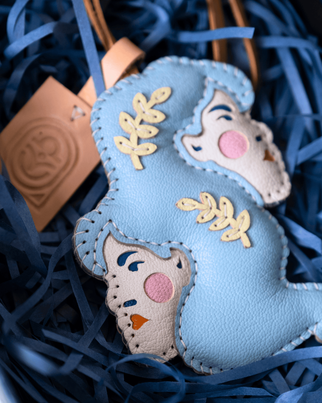 A light blue and cream leather ornament shaped like a mother and child, decorated with gold leaf leaf motifs and pink embroidered cheeks, nestled in blue paper shred.