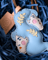 A light blue and cream leather ornament shaped like a mother and child, decorated with gold leaf leaf motifs and pink embroidered cheeks, nestled in blue paper shred.