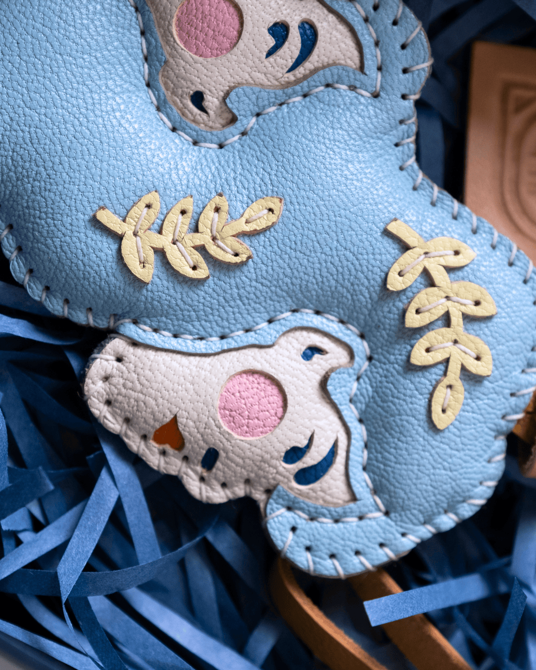 A close-up of a light blue textured leather patch featuring embroidered details, including a white bird with a pink circle, blue curved lines, and beige leaf motifs, resting on blue shredded paper.