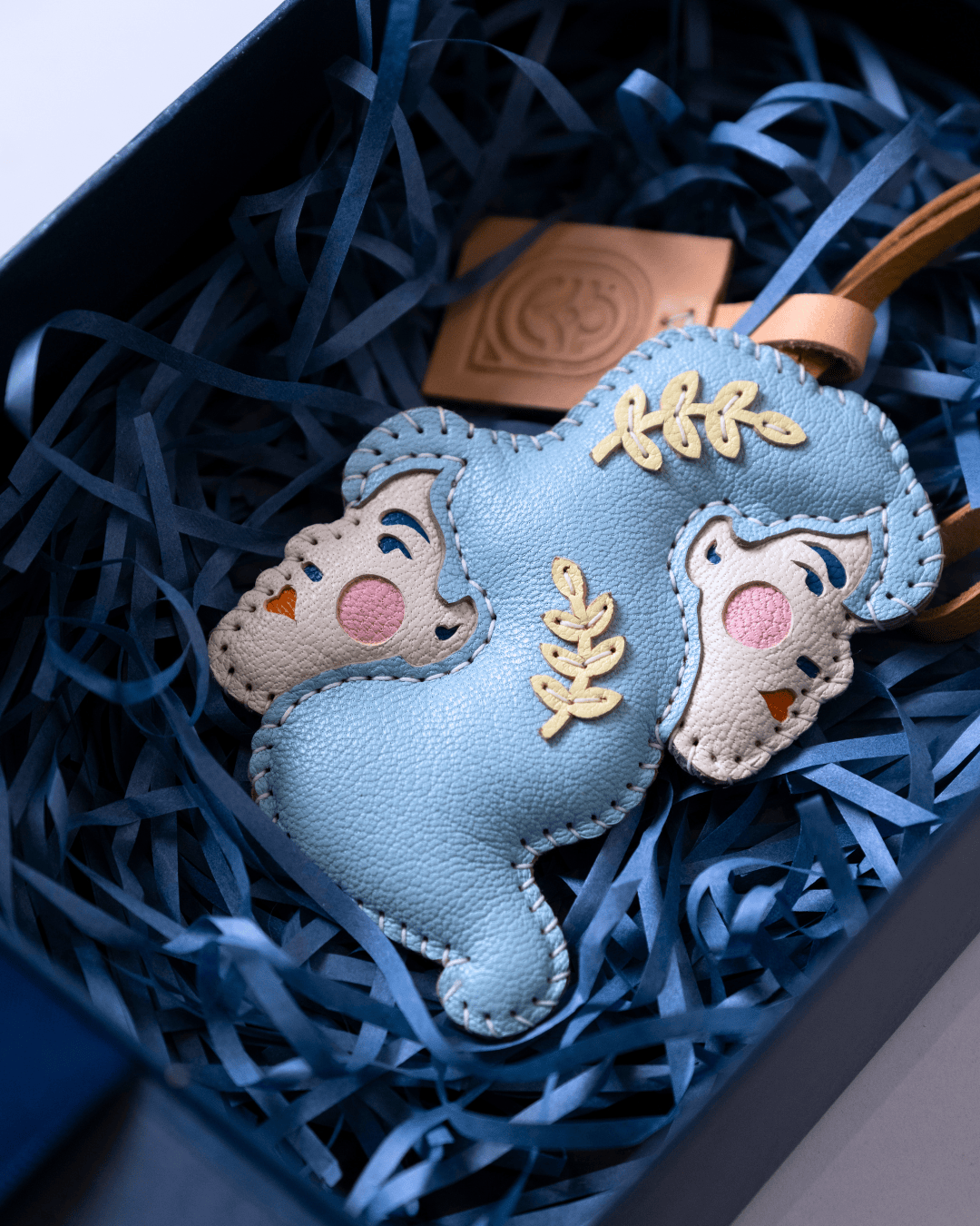 A light blue leather keychain charm shaped like a sleeping cartoon character, displayed inside a dark blue gift box filled with blue paper shred. The charm features gold-colored embroidered details and a small tan leather tag with an embossed logo.