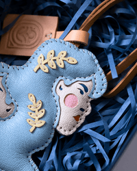 A close-up of a light blue leather horse-shaped bag charm or pouch with embroidered details, including a white horse face patch with a pink nose and blue stitched accents, placed among blue shredded paper and tan ribbon.