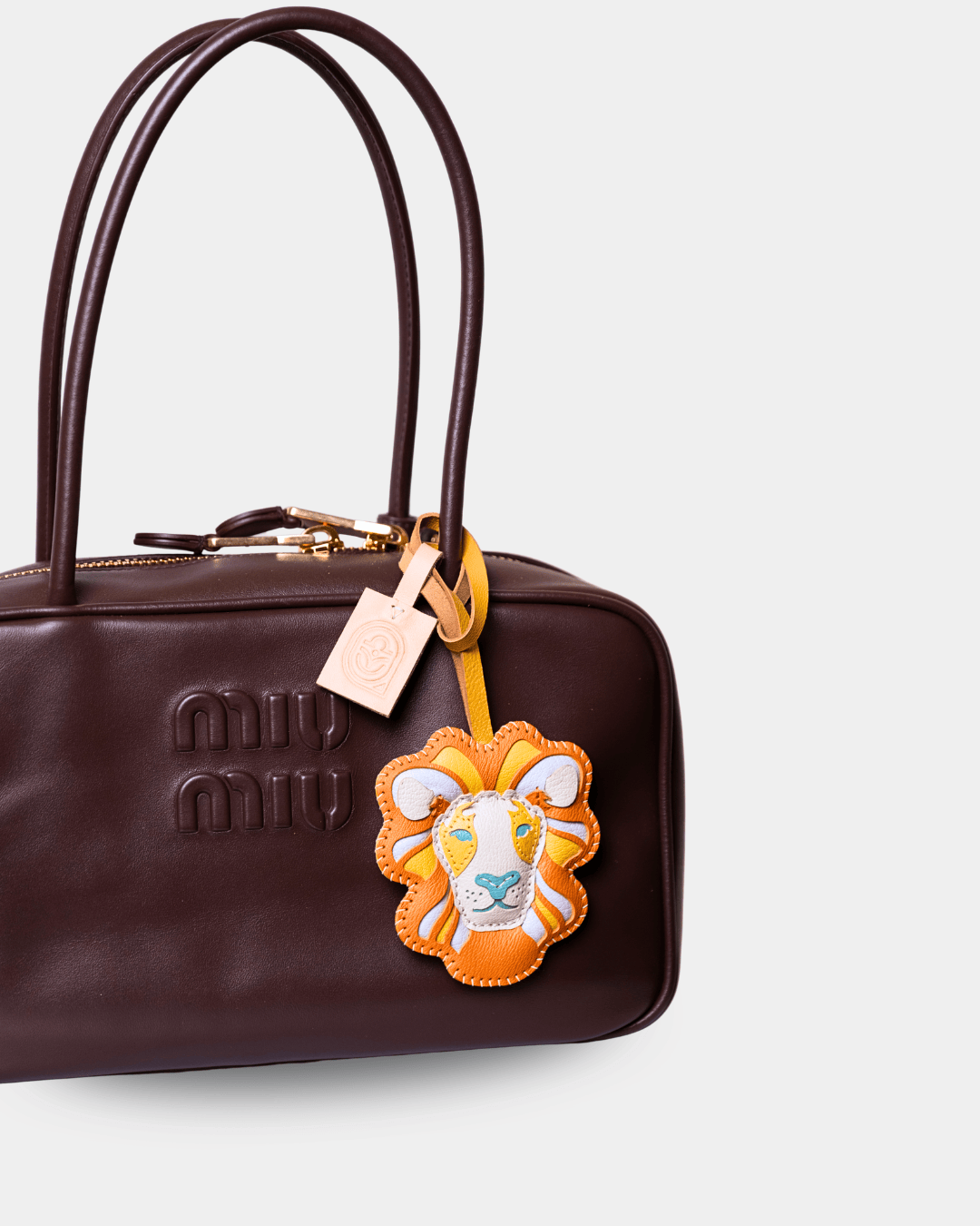 A dark brown leather Miu Miu handbag with a gold-tone zipper, featuring an embossed logo on the front, dual top handles, and a colorful lion-face leather charm.