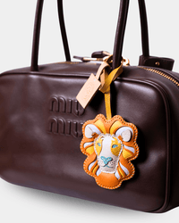 A close-up of a dark brown leather Miu Miu handbag featuring embossed logo lettering and a decorative lion-head luggage tag hanging from the zipper.