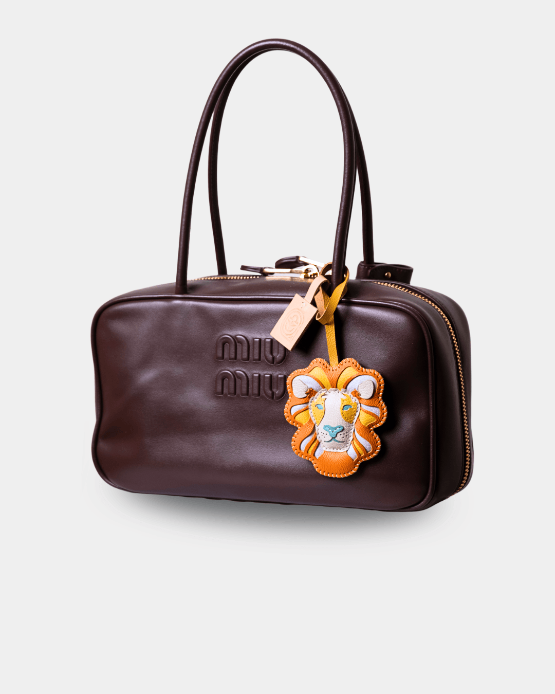 A dark brown leather Miu Miu handbag with a top handle, gold-tone zipper, and a lion patch charm, shown on a light gray background.